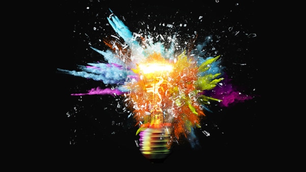 Creative light bulb explodes with colorful paint splashes and shards of glass on a black background.